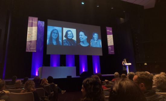 the blue lit stage at IATEFL, with a speaker behind a podium and four other speakers showing on the large projector before the seated audience.
