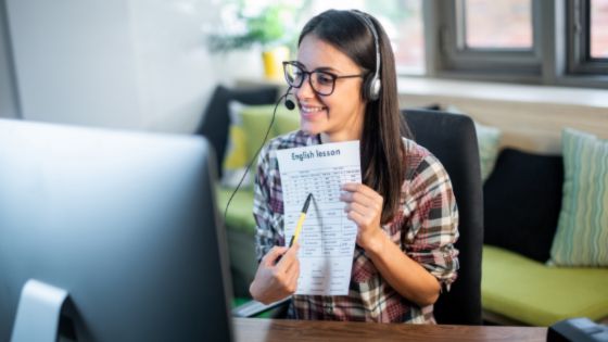 A woman teaches English on her computer while holding up a worksheet.