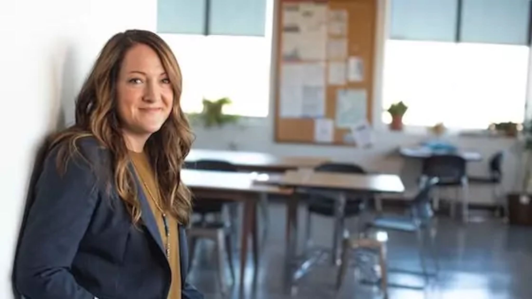 a teacher leaning against a wall smiling in an empty classroom.