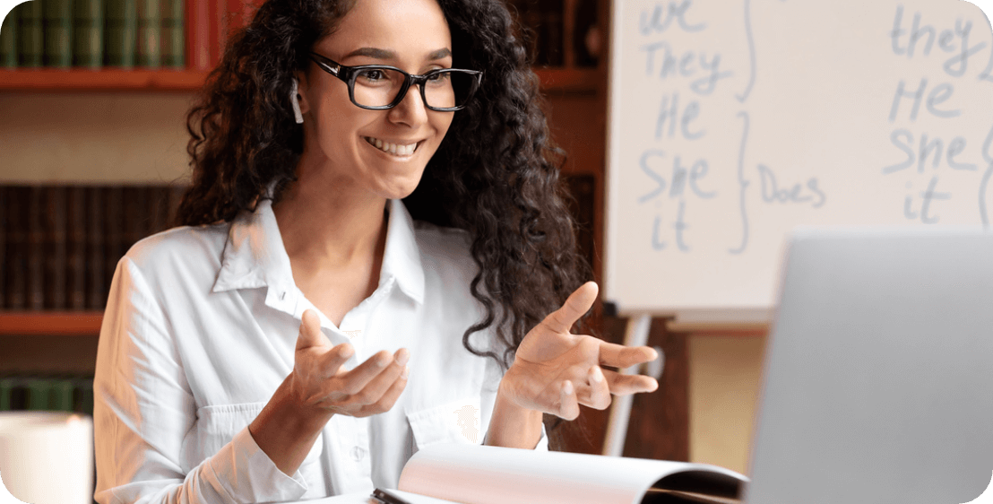 Meet some of our teachers A woman with long, curly hair and glasses smiles while looking at a laptop. She is wearing a white shirt and gesturing with her hands. Behind her, there's a whiteboard with English grammar notes, including pronouns and verbs.