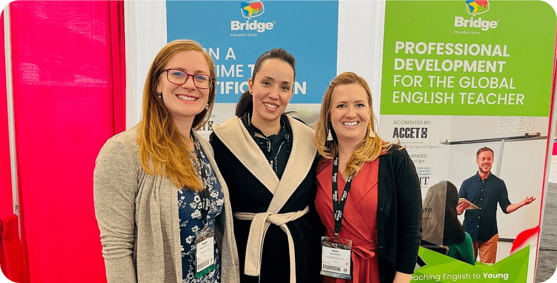 Certified Global English Teachers Three smiling women are standing in front of a booth display. The booth features banners with the text "Bridge Education Group" and "Professional Development for the Global English Teacher." The women are wearing conference lanyards and badges.
